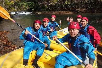 北海道のラフティングツアーでボート上に座り、ピースサインをする参加者たち。紅葉に囲まれた秋のアウトドア体験。